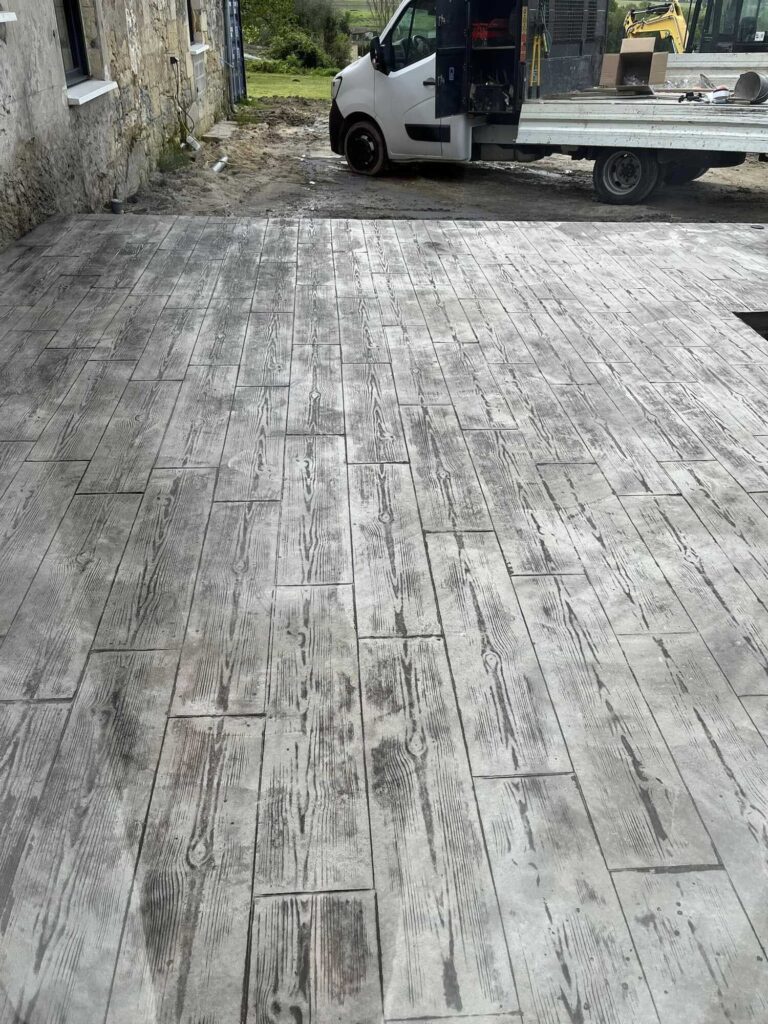 Béton texturé imitation bois : Patio extérieur moderne Dalle de béton gris texturée imitation bois, formant un patio extérieur devant un bâtiment en pierre et un camion de chantier.