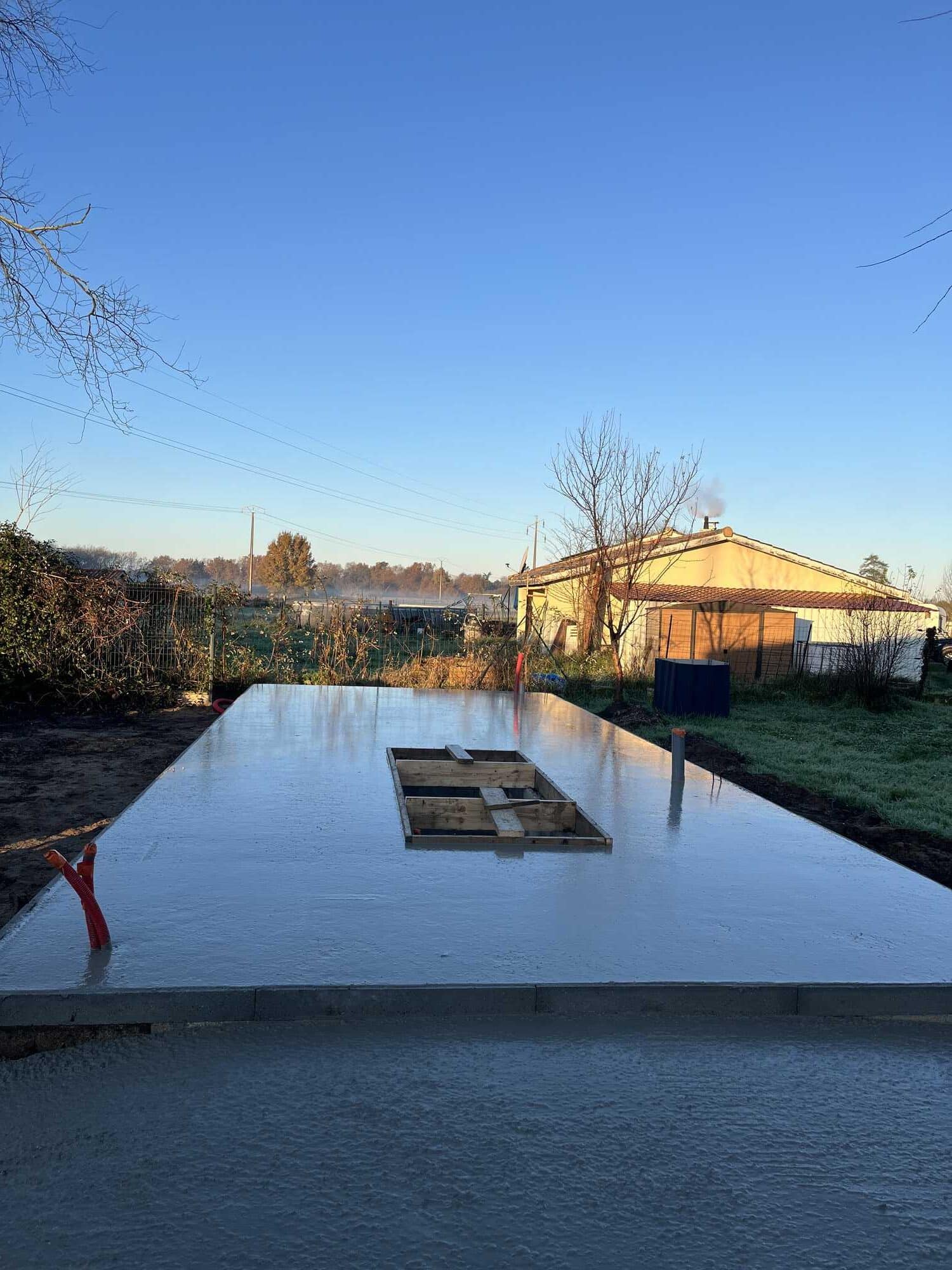 Dalle de béton fraîchement coulée pour une fondation, avec ouverture et conduits visibles. Ciel bleu matinal sur un chantier en campagne.
