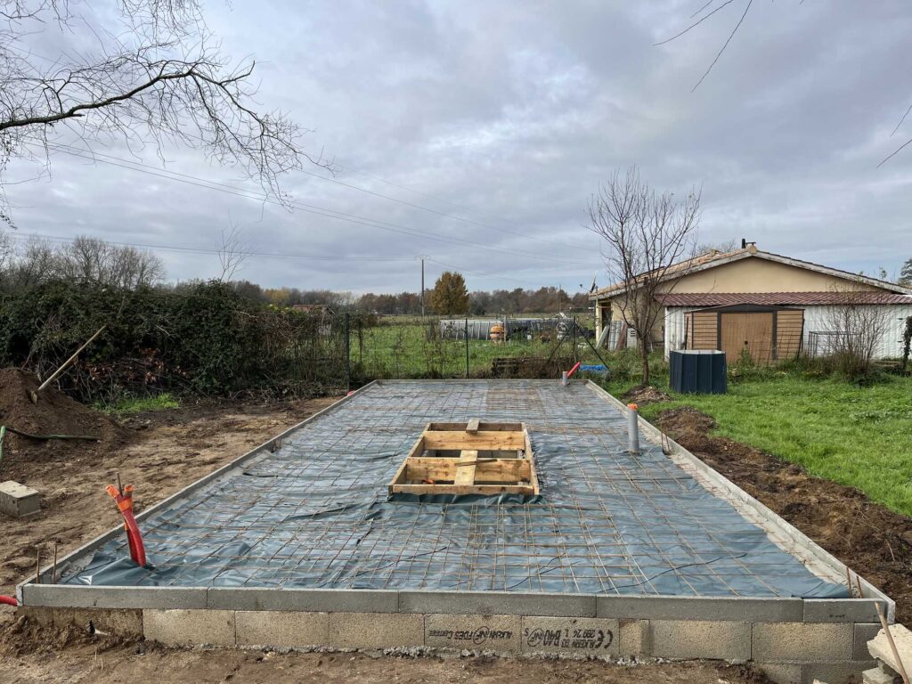 Fondations Maison Individuelle : Préparation Dalle Béton Fondations de maison avec treillis d'armature sur membrane et blocs béton ALCEN. Forme bois centrale et conduits orange.