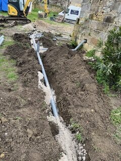 Installation de drainage: tranchée, tuyaux PVC et travaux Longue tranchée creusée dans la terre avec des tuyaux PVC gris pour une installation de drainage. Une évacuation part du mur en pierre à droite. Une excavatrice est visible à gauche.