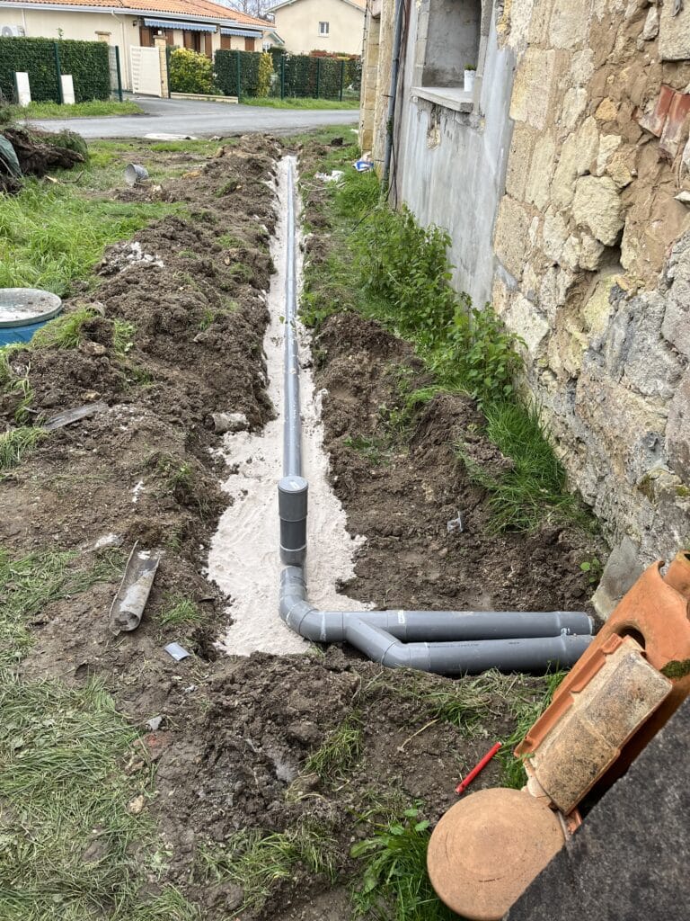 Installation tuyaux de drainage PVC: plomberie maison en cours Tranchée ouverte avec tuyaux de plomberie gris installés pour drainage, entourés de sable, devant une maison en pierre.