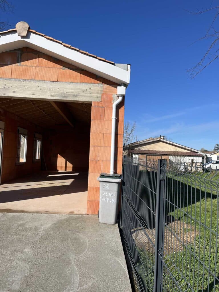 Maison Neuve: Construction Briques, Avancement Chantier Maison neuve en briques oranges, façade ouverte, sous ciel bleu. Gouttière blanche, poubelle '26 DENIS GAPA', clôture noire.