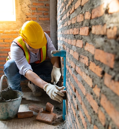Ouvrier en bâtiment avec casque et gilet de sécurité installant une conduite d'eau bleue dans un mur de briques rouges sur un chantier.