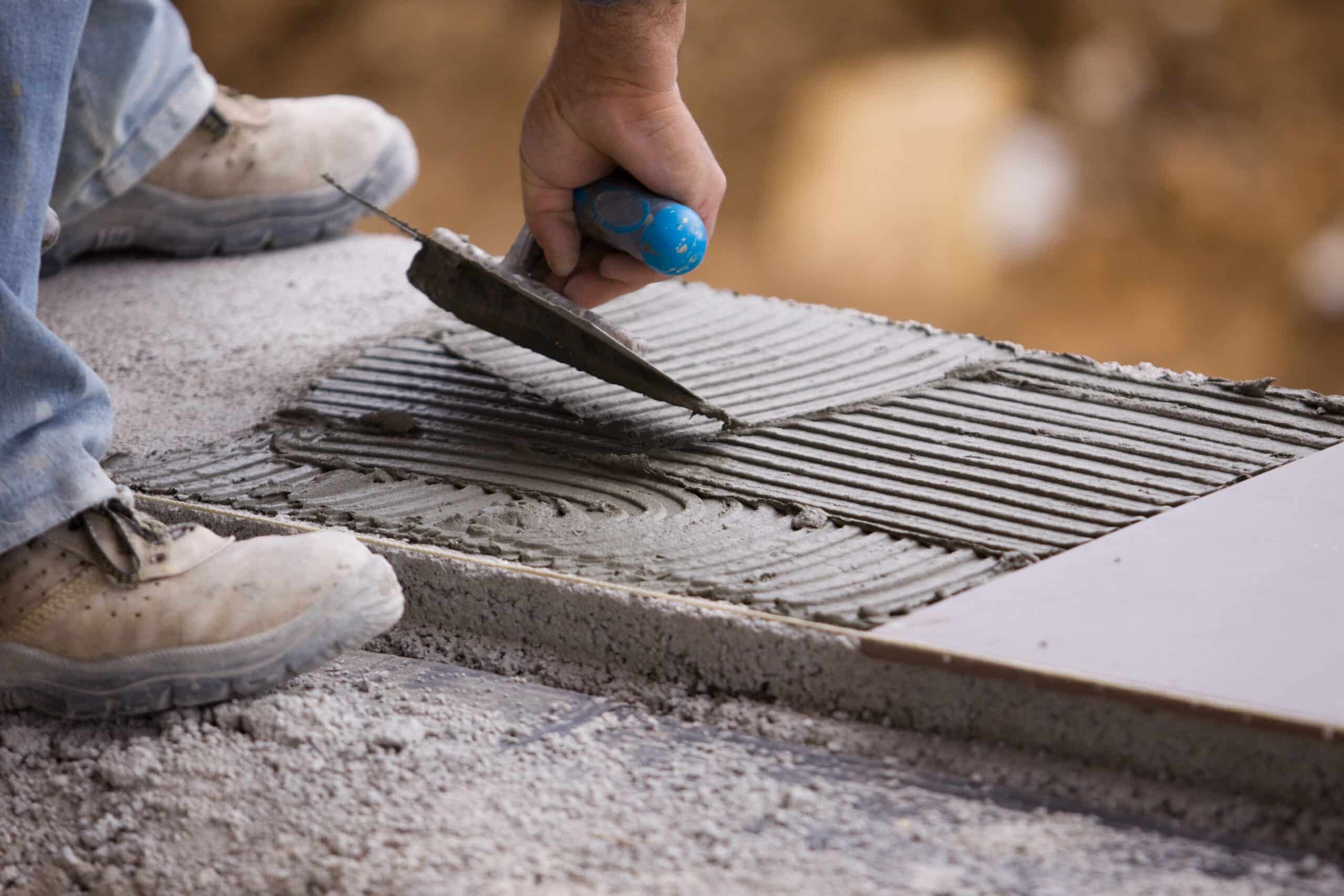 Maçon appliquant du mortier adhésif avec une truelle dentelée sur une surface en béton pour la pose de carrelage, pieds en bottes de travail.