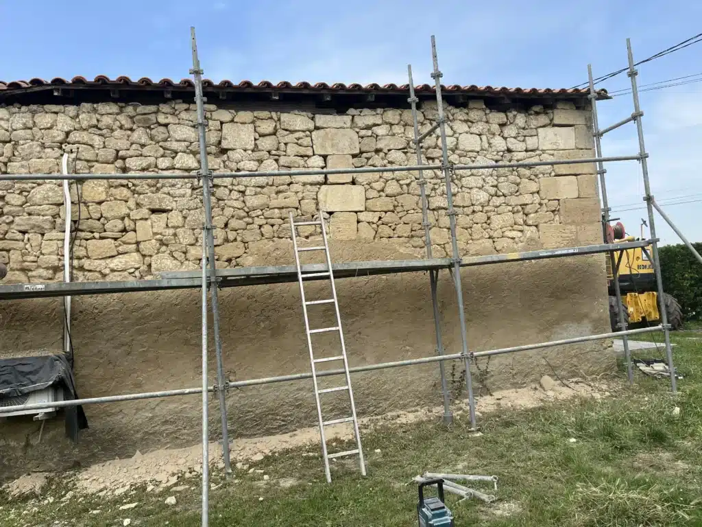 Rénovation mur pierre : échafaudage et engins de chantier Mur en pierre et terre d'une maison en rénovation avec échafaudage ACGMABI, échelle, et excavatrice Komatsu jaune. Ciel bleu.