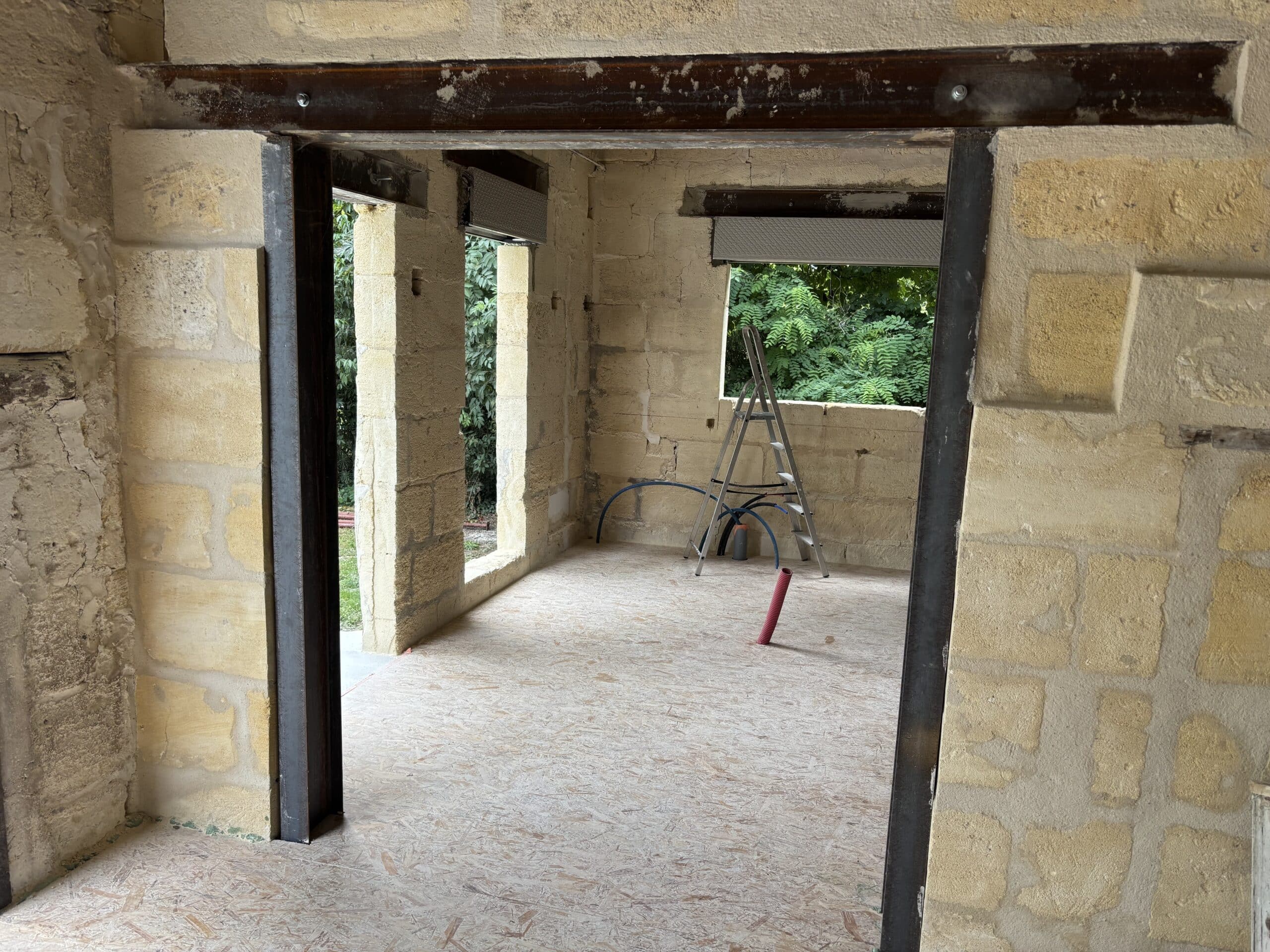 Chantier de rénovation avec linteau métallique, murs en pierre apparente, sol OSB et conduits techniques au sol.