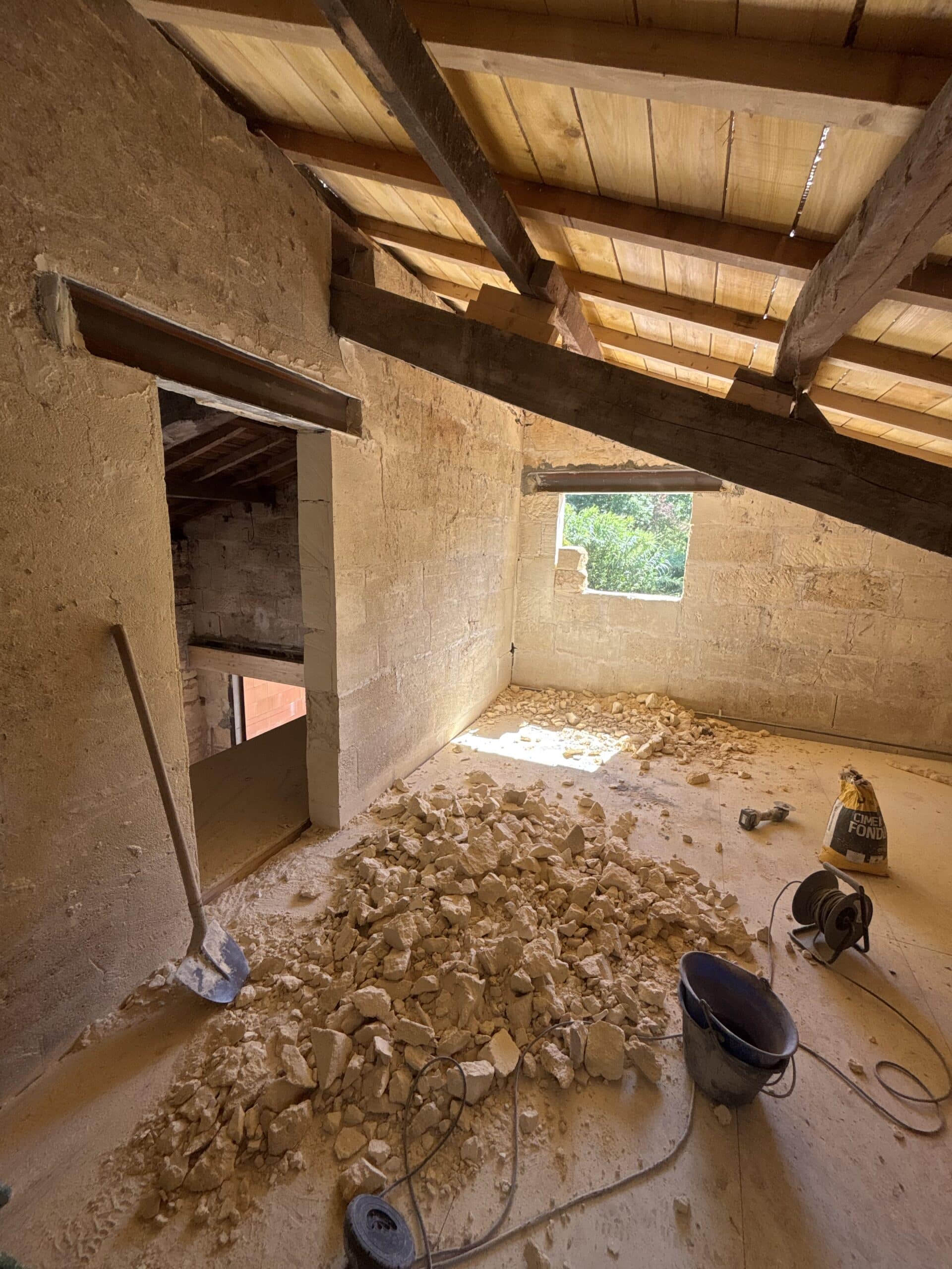 Chantier de rénovation d'un grenier avec murs en pierres, poutres apparentes, gravats au sol et outils de maçonnerie.