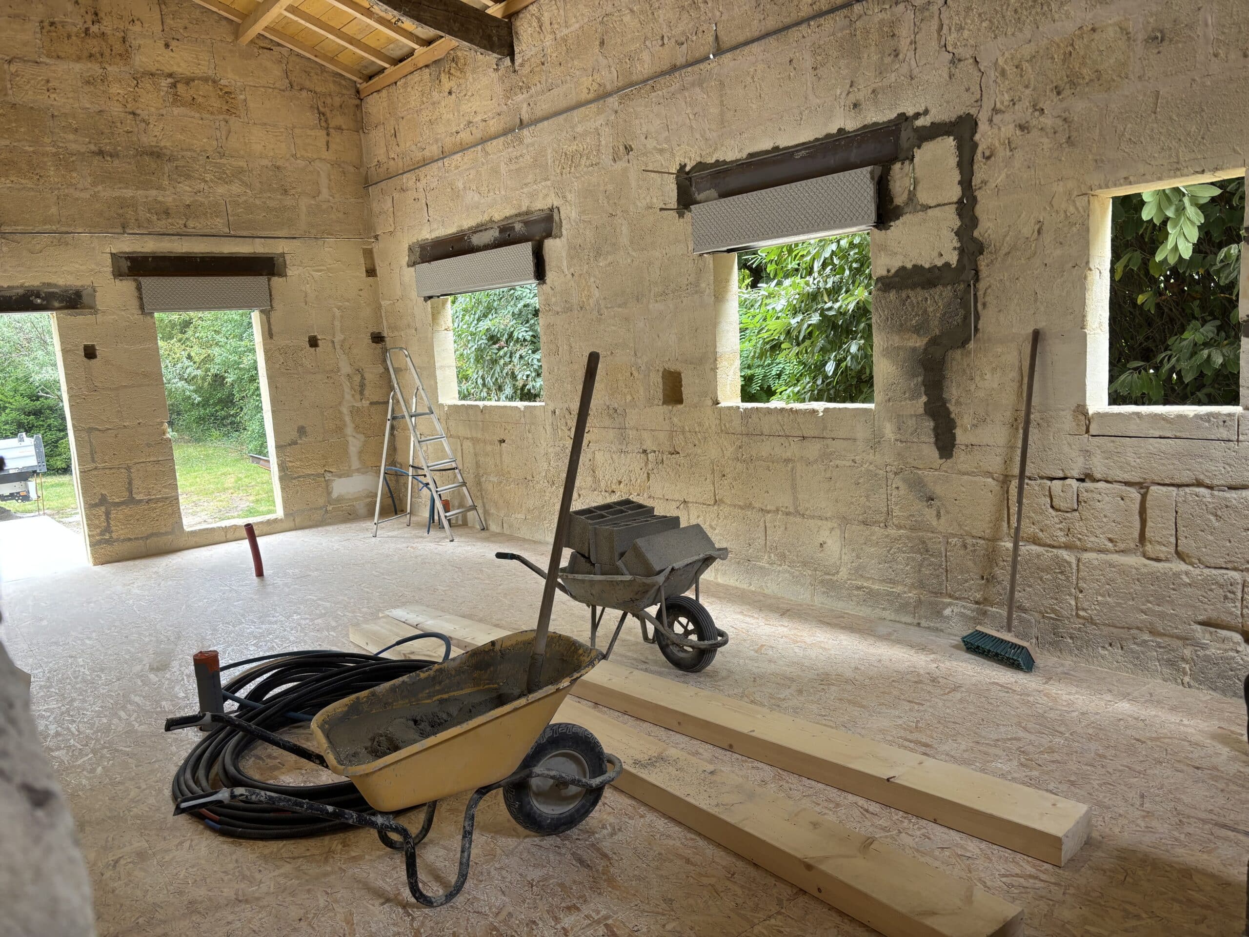 Chantier de rénovation intérieure : murs en pierre de taille, brouettes, matériaux de maçonnerie et sol OSB.
