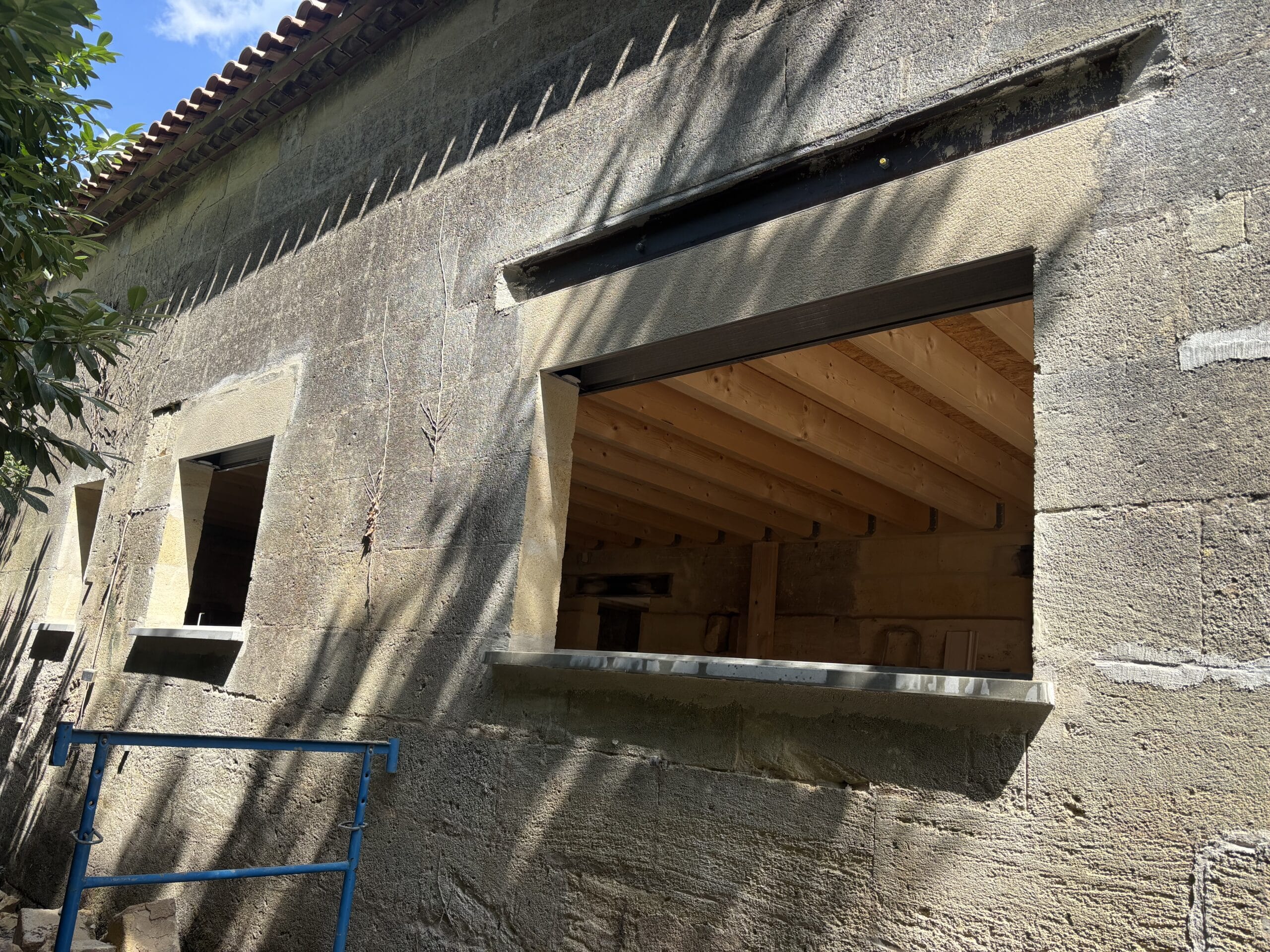Mur en pierre de taille en rénovation avec ouvertures de fenêtres révélant une charpente bois et linteau métallique.